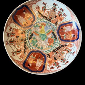 Puede incluir: Un plato de cerámica redondo con un medallón central que muestra un fénix verde y amarillo rodeado de motivos florales. El borde presenta paneles alternos de barcos de vela y figuras con vestimenta tradicional, en rojo, azul y dorado.