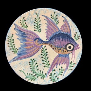 May include: A hand-painted ceramic plate featuring a stylized fish design. The fish is primarily purple and blue with orange accents, set against a white background with green and red plant-like motifs. The plate has a blue rim.