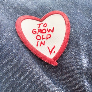 May include: Red heart-shaped patch with white fabric in the center. The text "TO GROW OLD IN V." is embroidered in red on the white fabric.