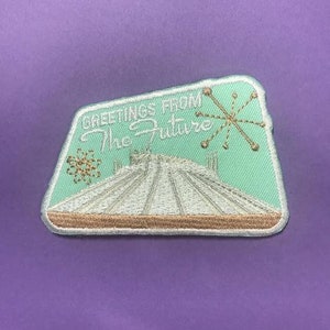 May include: A trapezoidal embroidered patch with a light mint green background. It features white stitching of a conical structure, gold starburst and atom icons, and the text "GREETINGS FROM The Future" in white and gold thread.