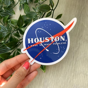 May include: A round sticker with a blue background and a white circle design. The sticker has the text "HOUSTON, I HAVE SO MANY PROBLEMS" in white. A red swoosh crosses the circle. The sticker is held by a hand.