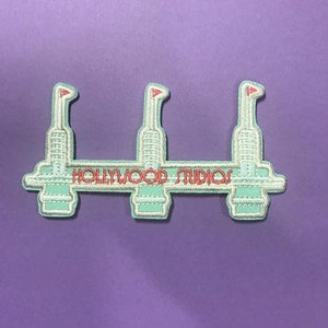 May include: A light blue embroidered patch with three white towers and the text "HOLLYWOOD STUDIOS" in red. The towers have red flags on top.