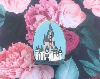 Può includere: Uno spillo in smalto blu e bianco con un'illustrazione di cartone animato del castello di Cenerentola con il testo "castle dreaming" in lettere dorate.