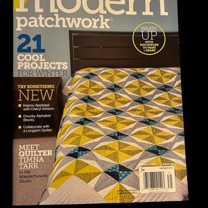 May include: A magazine cover for Modern Patchwork featuring 21 cool quilting projects for winter. The cover features a quilt with a geometric pattern in shades of yellow, gray, and teal. The text "Warm Up with Patchwork to Make + Share" is featured on the cover.