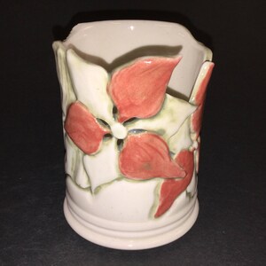 May include: A white ceramic vase with a floral design. The vase has a textured surface and features red and green flowers.