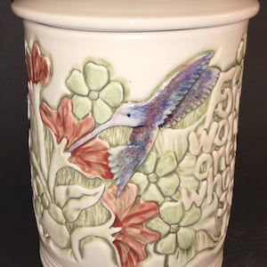 May include: White ceramic jar with a lid, featuring a 3D design of a hummingbird in flight, surrounded by red and green flowers. The jar has the words "Five words are what" written on the side.