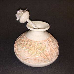 May include: A white ceramic vase with a carved floral design in shades of pink and green. The vase has a round body and a small opening at the top. A white wick is inserted into the opening.