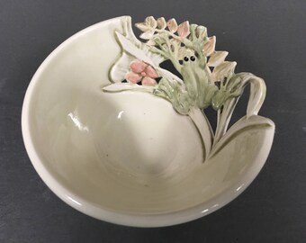 May include: A white ceramic bowl with a green frog and pink flowers sculpted on the side. The bowl is designed with a raised rim and a scalloped edge.