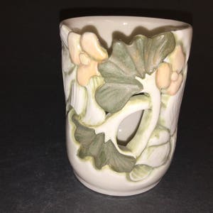 May include: A white ceramic cup with a floral design. The cup features raised green leaves and pink flowers.