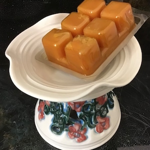 May include: A set of six amber wax melts in a clear plastic container, sitting on a white ceramic pedestal with a floral design.