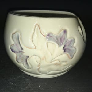 May include: A white ceramic yarn bowl with a floral design. The bowl has a hole on the side for the yarn to pass through. The flowers are painted in shades of purple and pink.