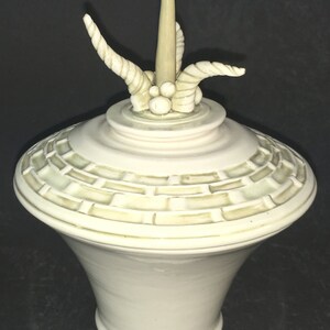 May include: A white ceramic jar with a lid and a decorative top. The top features a tall, pointed spire and two curved, twisted horns. The jar has a textured, grid-like pattern around the top.