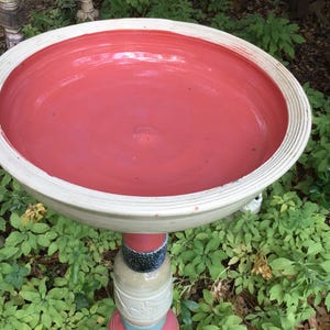 May include: A handcrafted bird bath with a coral-colored basin and a white rim. The pedestal is composed of stacked, decorative ceramic elements in various colors. The bird bath is surrounded by green foliage.