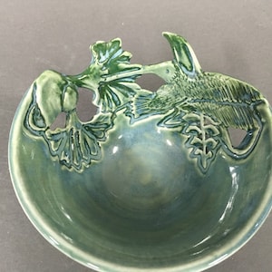 May include: A green ceramic bowl with a leafy design. The bowl has a cutout design of leaves and a bird.