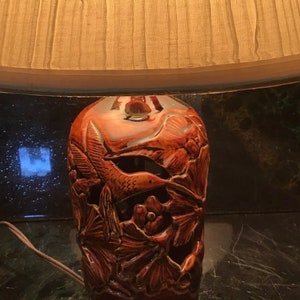 May include: A brown ceramic table lamp with a white lampshade. The lamp base features a detailed design of hummingbirds and flowers.