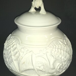 May include: White ceramic jar with a lid and a sculpted floral design. The jar has a handle on the lid.