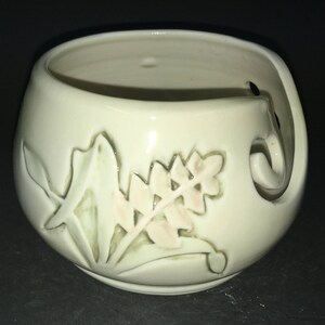 May include: White ceramic yarn bowl with a leaf design. The bowl has a hole on the side for yarn to pass through.