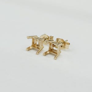 May include: A pair of gold-colored stud earrings. Each earring features a small, open-frame design resembling a miniature chair. The earrings are set against a plain white background, highlighting their intricate details and warm metallic hue.