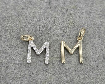 May include: Two letter M pendants. One pendant is gold-toned with a row of small clear stones embedded along the surface. The second pendant is a plain, smooth gold-toned metal. Both pendants feature a small circular loop at the top for attachment.