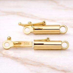 May include: Two gold-colored jewelry clasps, one open and one closed, are displayed against a marble background. The clasps have a cylindrical design with a lever closure and a small loop at each end. The text "14K" is visible on one clasp.