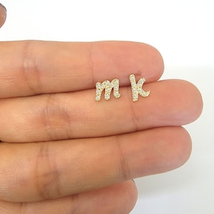 Puede incluir: Un par de pendientes de botón dorados, cada uno con la forma de una letra minúscula. La letra "m" y la letra "k" están incrustadas con pequeñas gemas transparentes. Los pendientes se muestran sobre un fondo claro.