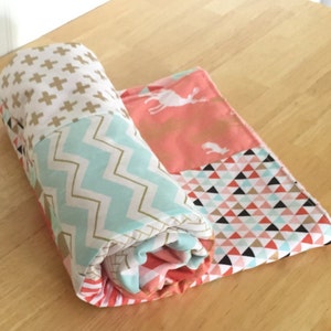 Puede incluir: Una colcha de retazos enrollada con una variedad de patrones, incluyendo cruces doradas, chevron turquesa, coral con ciervos blancos y triángulos rosas y negros.