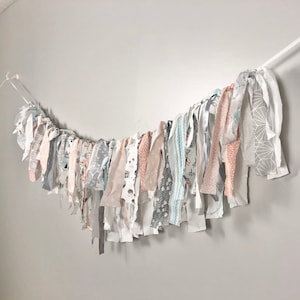 Puede incluir: Una guirnalda de tela blanca, gris y melocotón con una variedad de patrones. La guirnalda está enhebrada en un cordón blanco y cuelga de una pared.