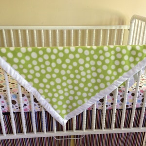 May include: A white crib with a green and white polka dot blanket draped over the top. The crib has a white mattress with a colorful patterned sheet.