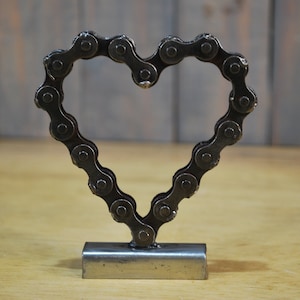 May include: A metal heart sculpture made from a bicycle chain. The heart is standing on a metal base.