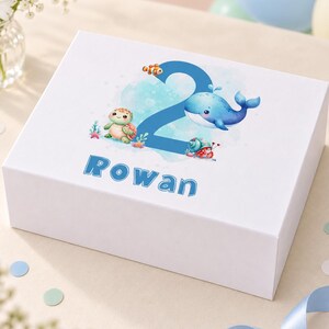 May include: A white rectangular gift box featuring a large blue number two, the name Rowan in blue letters, and illustrations of a whale, a sea turtle, a hermit crab, and a clownfish surrounded by blue watercolor accents.