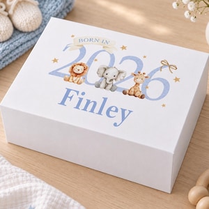 May include: A white rectangular keepsake box featuring the text BORN IN 2026 and the name Finley in blue. The design includes illustrations of a lion, an elephant, and a giraffe, accented with small gold stars and a gold bow graphic.