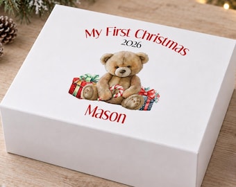 Puede incluir: Una caja de regalo rectangular blanca con una ilustración impresa de un oso de peluche marrón que sostiene un bastón de caramelo, rodeado de regalos. El texto dice My First Christmas 2026 y el nombre Mason en letras rojas.