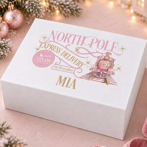 May include: A white rectangular gift box featuring a pink and gold graphic of a train and the text NORTH POLE EXPRESS DELIVERY, LAPLAND, 25th December, and the name MIA printed in gold on the front surface of the box.