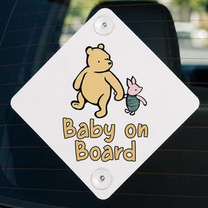 Op de afbeelding: Een ruitvormig wit autobord met een gele beer die de hand vasthoudt van een klein roze biggetje. Onder de figuren staat de tekst Baby on Board in gele letters met een zwarte omlijning. Het bord is voorzien van twee doorzichtige zuignappen.