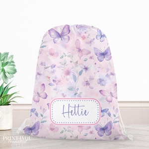 May include: A drawstring backpack featuring a light pink background with a watercolour pattern of purple butterflies, pink flowers, and green leaves. A central white rectangular label with a pink border displays the name Hettie in purple script.