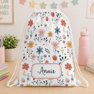 May include: White drawstring bag featuring a vibrant floral design with red, pink, yellow, and blue flowers. The name "Annie" is displayed within a decorative frame. The bag has white drawstrings.