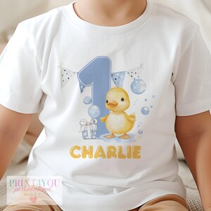 A white baby t-shirt featuring a cute yellow duckling next to a large blue number "1", with the name "Charlie" printed below. The design includes a gift box and festive bunting.