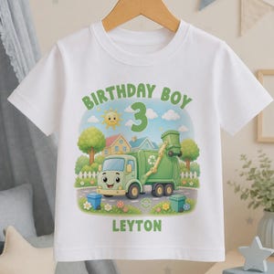 May include: A white short-sleeve t-shirt featuring a graphic of a green cartoon refuse truck, a smiling sun, houses, and trees. The text reads BIRTHDAY BOY, the number 3, and the name LEYTON in green letters. The design includes blue and green bins.