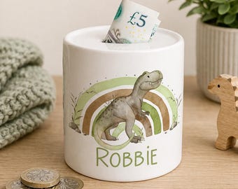 May include: A white ceramic money box featuring a watercolour illustration of a green dinosaur standing in front of a brown and green rainbow. The name Robbie is printed in green letters below the dinosaur. A five-pound note is inserted in the top slot.