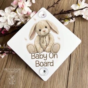 May include: White diamond-shaped "Baby On Board" sign with a plush bunny illustration. The bunny is beige with a cream-colored bow. The sign has two suction cups for attachment.
