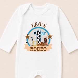 A white onesie with a graphic design featuring a cow, a horse, and a cowboy hat, along with the text "LEO'S 1ST RODEO".