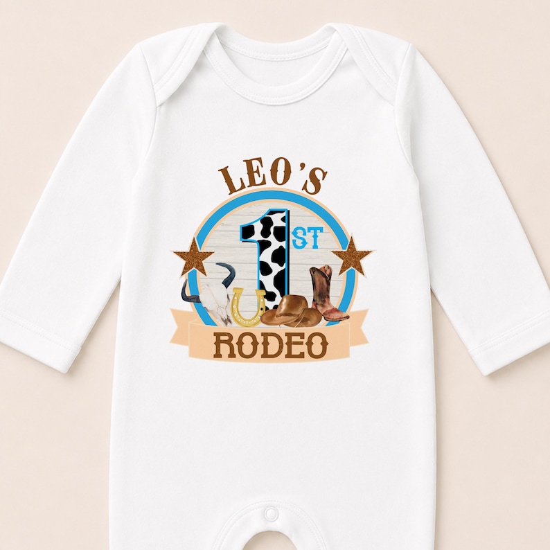 A white onesie with a graphic design featuring a cow, a horse, and a cowboy hat, along with the text "LEO'S 1ST RODEO".