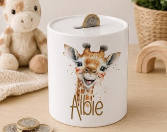 May include: A white ceramic money box featuring a watercolor illustration of a smiling giraffe with brown spots and the name Albie printed in brown script below. A single metallic coin is inserted into the slot at the top of the cylinder.