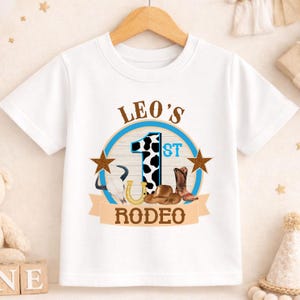 A white t-shirt with a graphic design featuring a cow, a horseshoe, and the text "LEO'S 1ST RODEO" printed on it.