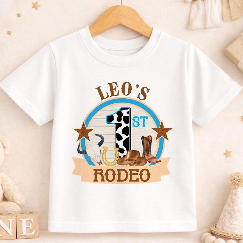 A white t-shirt with a graphic design featuring a cow, a horseshoe, and the text "LEO'S 1ST RODEO" printed on it.