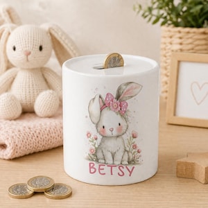 May include: A white ceramic cylindrical money box featuring a watercolour illustration of a small rabbit with a pink flower bow on its head, surrounded by pink flowers. The name BETSY is printed in pink capital letters below the rabbit illustration.