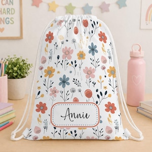 May include: A white drawstring backpack featuring a repeating pattern of small flowers in shades of orange, pink, blue, and yellow. The bag has a rectangular label on the front with a dotted border containing the name Annie in black script.