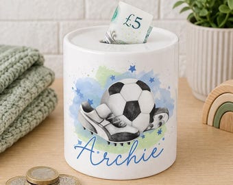 May include: A white ceramic money box featuring a watercolour illustration of a football and a boot. The name Archie is printed in blue script below the graphic. A five-pound note is inserted into the top slot of the cylindrical container.