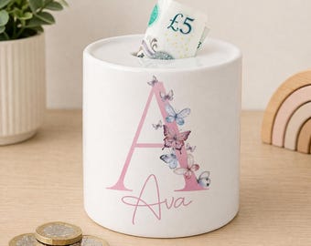 May include: A white ceramic money box featuring a large pink letter A decorated with several small blue and pink butterflies. The name Ava is printed in pink script below the letter. A five-pound note is inserted into the top slot of the container.