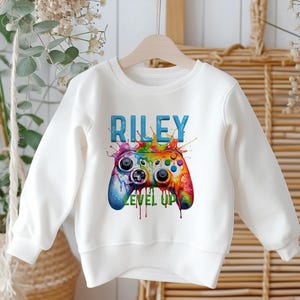 K&ouml;nnte beinhalten: Wei&szlig;es Sweatshirt mit einer farbenfrohen Videospiel-Controller-Grafik und dem Namen "RILEY" dar&uuml;ber. Der Controller ist eine lebendige Mischung aus Farben, mit den Worten "LEVEL UP" darunter. Das Design hat einen Splash-Art-Stil.